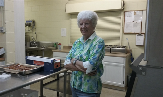 An elderly woman stands in a well-lit kitchen, smiling brightly while preparing meals.