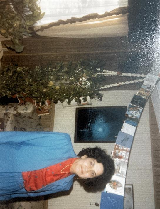 A woman stands confidently in a blue suit with a red scarf, smiling indoors by a decorated wall.