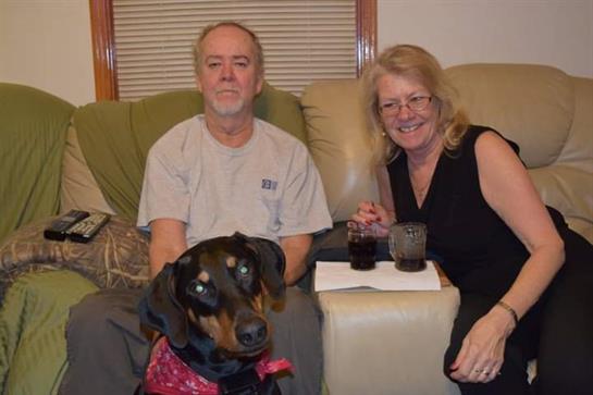 A couple relaxes on a sofa holding drinks while their dog sits happily in front of them.