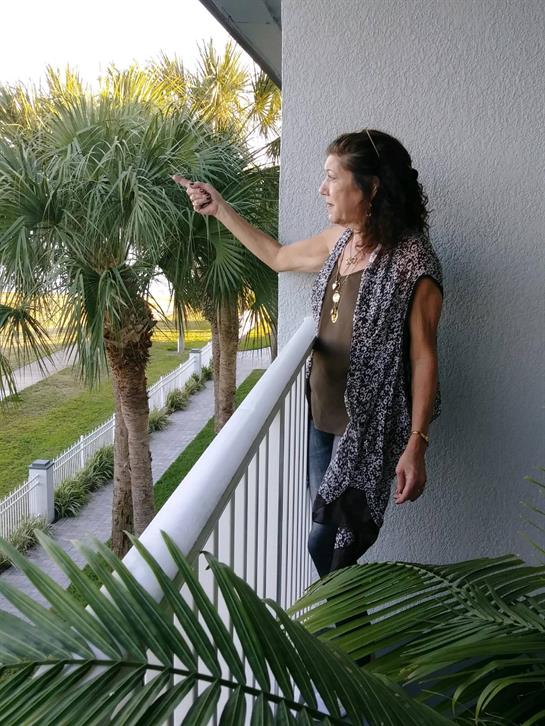 A woman stands on a balcony, happily pointing at palm trees under bright sunlight.