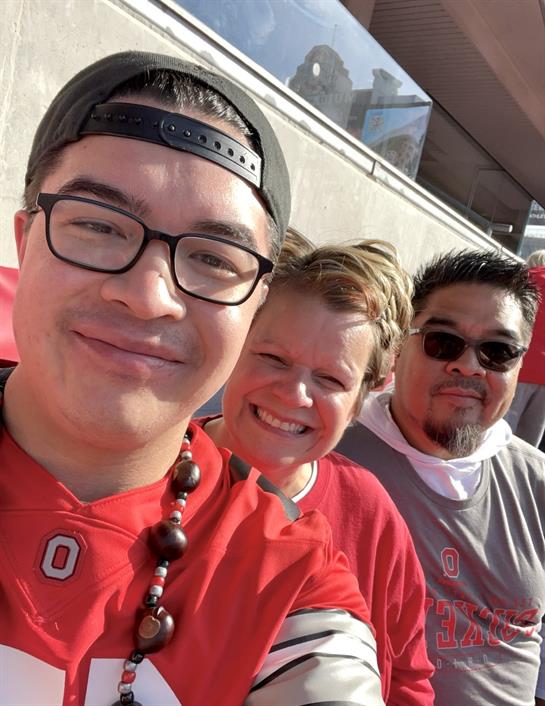 Three fans wear team colors, smiling and celebrating during a vibrant football game at the stadium.