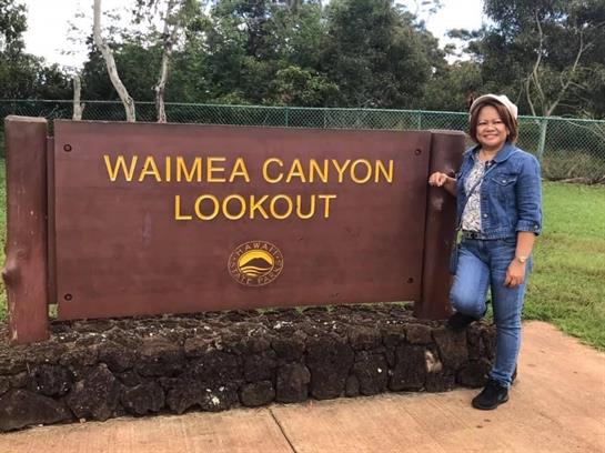 A woman stands beside the Waimea Canyon Lookout sign, enjoying the beautiful scenery.