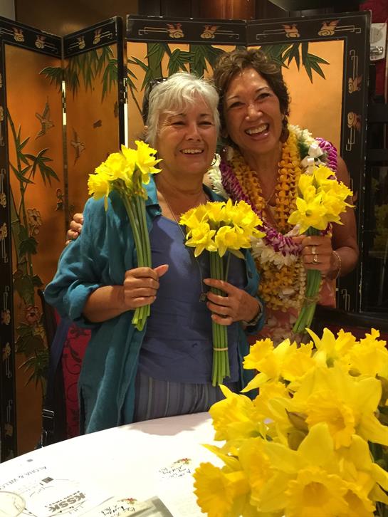 Two women smile with vibrant yellow flowers in a festive indoor celebration.