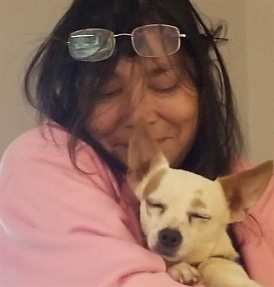 A woman wearing glasses smiles warmly as she cuddles her small dog in a relaxed indoor space.