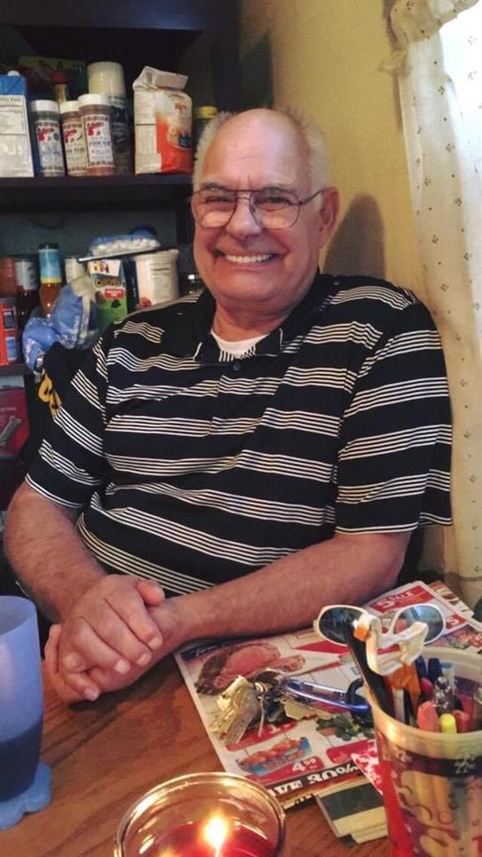 An older man in glasses smiles warmly in his cozy home, surrounded by keepsakes and food.