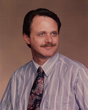 A man poses confidently in a studio, wearing a light striped shirt and a decorative tie, smiling.