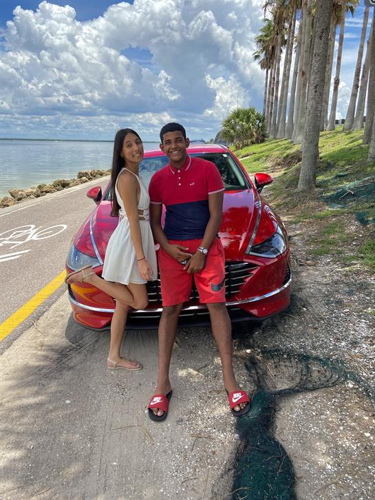 Two individuals stand smiling by a red vehicle along a coastal road under a sunny sky.