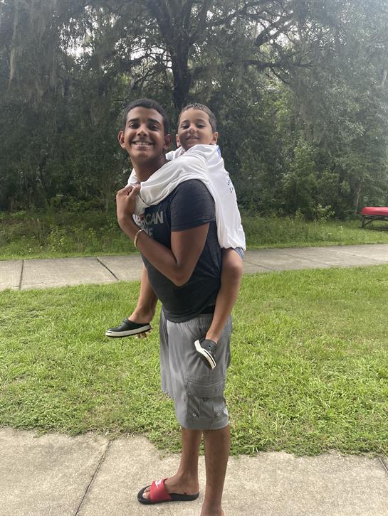 A boy happily carries his younger sibling on his back while standing in a grassy area.