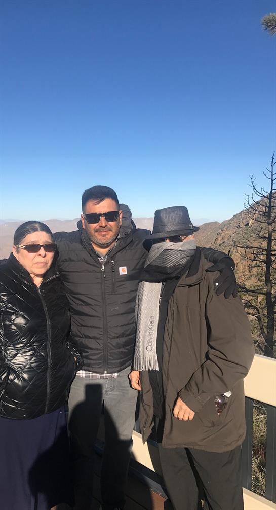 Three individuals pose together at a mountainous viewpoint, showcasing warm clothing and sunglasses.