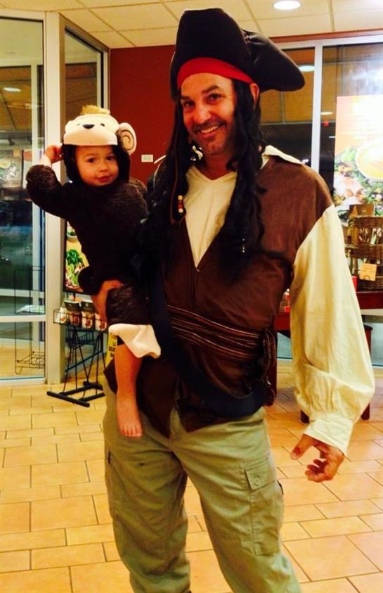 A child in a monkey costume is happily held by an adult in a pirate outfit at a store.