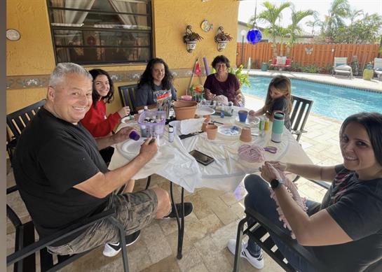 People are smiling and chatting at a table filled with food and drinks near a pool on a sunny day.