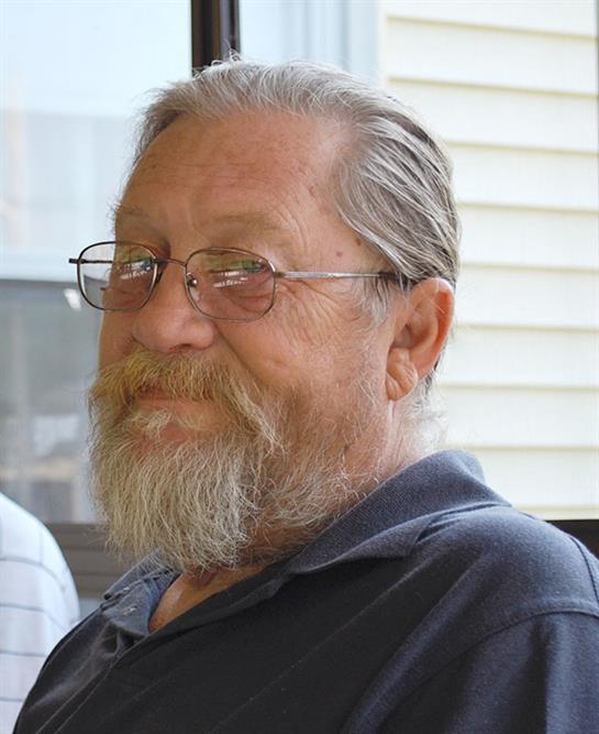 An elderly man sits indoors, looking friendly and enjoying a conversation.