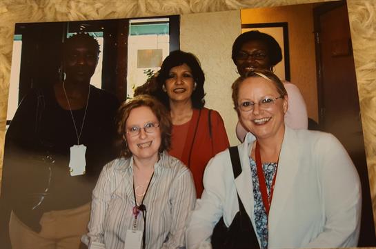 Five women stand together smiling inside a well-lit room, showing camaraderie and connection.