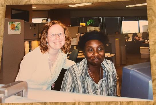 Two colleagues smile while posing together in a busy office environment full of workstations.