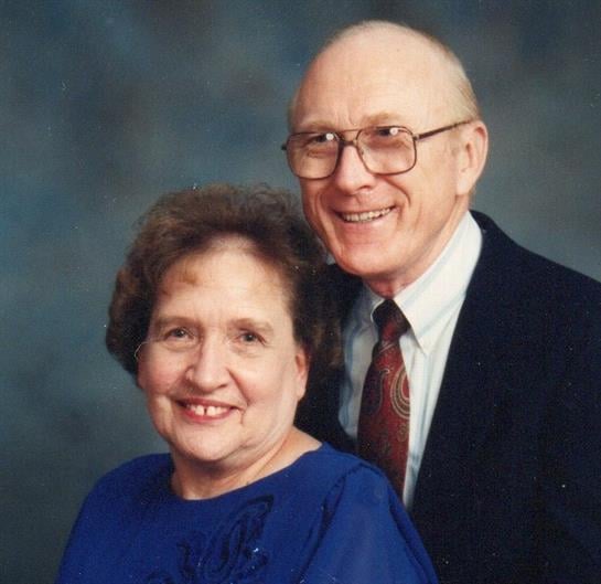 Elderly couple poses together, wearing formal clothes and smiling warmly in a studio setting.