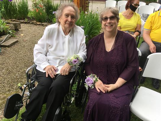 Two women sit together in wheelchairs at a garden wedding, smiling and enjoying the festivities.