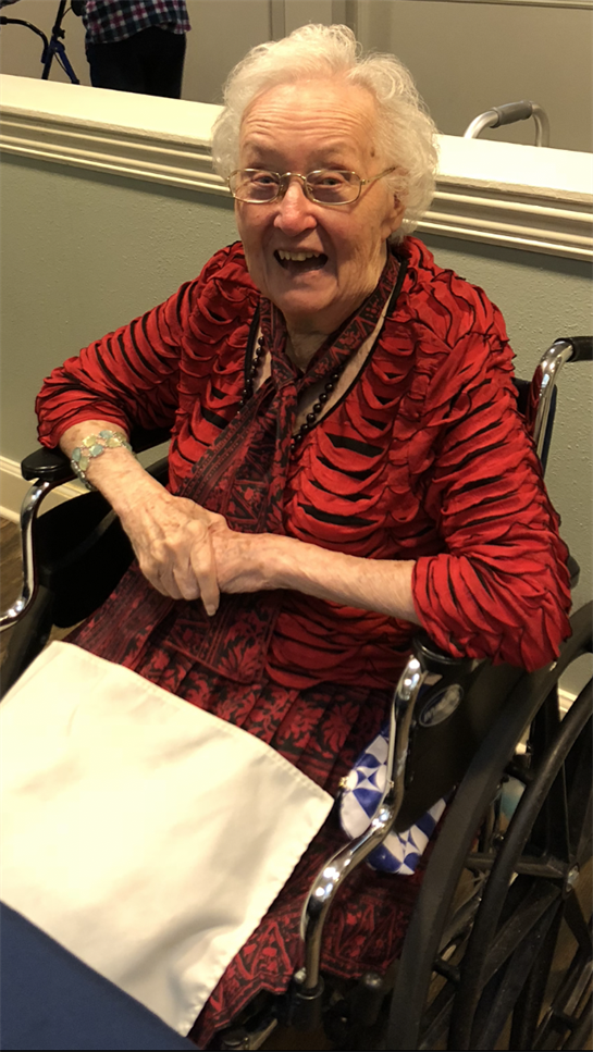 A joyful elderly woman wearing a red blouse sits in a wheelchair, interacting with others.