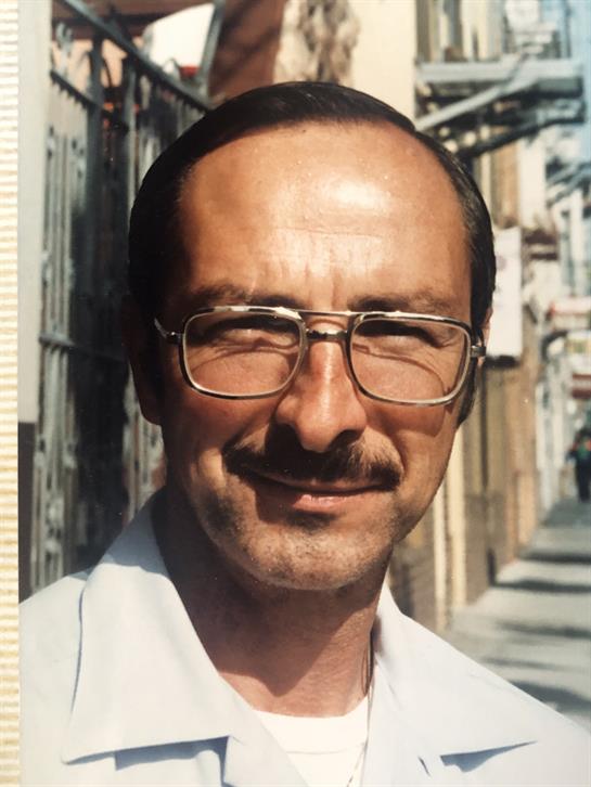 A man stands outdoors, smiling at the camera while wearing glasses in a vibrant street.