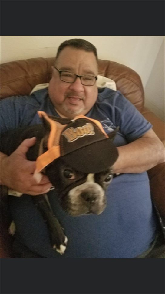 A man with glasses smiles while holding a French Bulldog wearing a cap in a cozy setting.