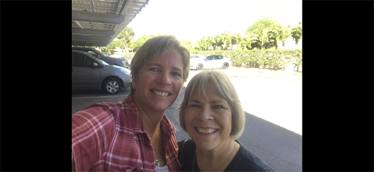 Two women pose happily together in a bright and sunny parking lot while smiling.