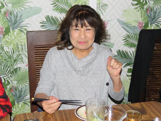 A woman is smiling and holding chopsticks while sitting at a table with a tropical decor.