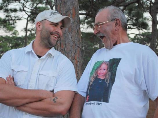 Two men smile and laugh together in a natural setting, embracing a special moment.