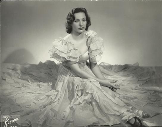 A woman sits elegantly wearing a vintage dress, showcasing classic fashion in a studio setting.