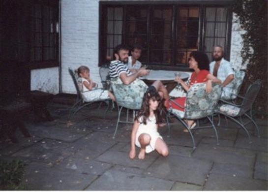 Children play on the ground while adults chat and relax outside a house on a sunny day.