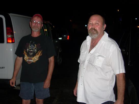 Two men are standing next to a white van, enjoying a relaxed moment at night.