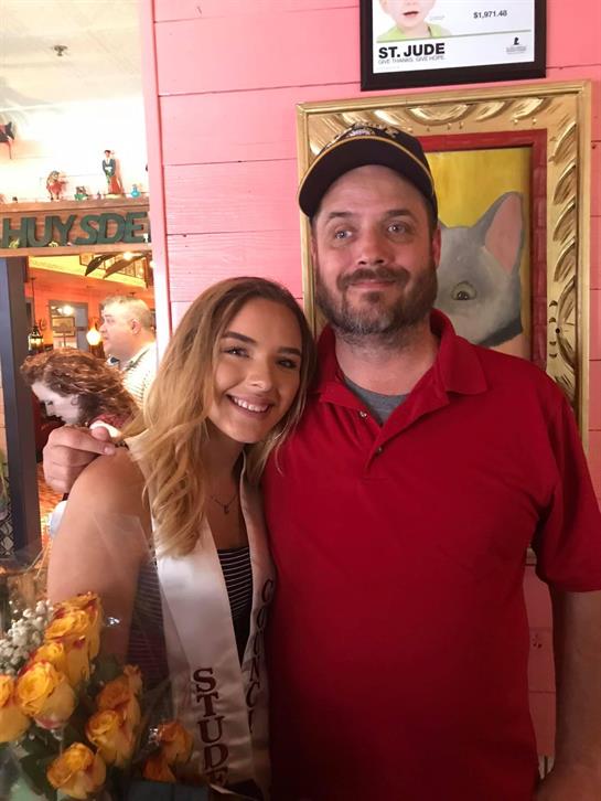 Young woman in a sash stands beside a man, both smiling, in a festive venue filled with decorations.