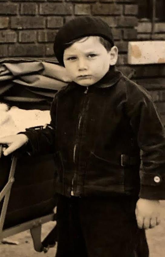 A young boy in a dark jacket holds a wooden cart, looking serious in a rustic environment.