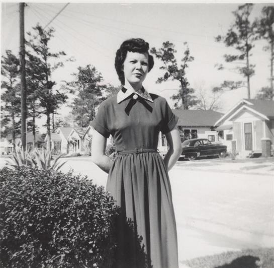 A woman in a vintage dress poses confidently beside green bushes on a sunny street.