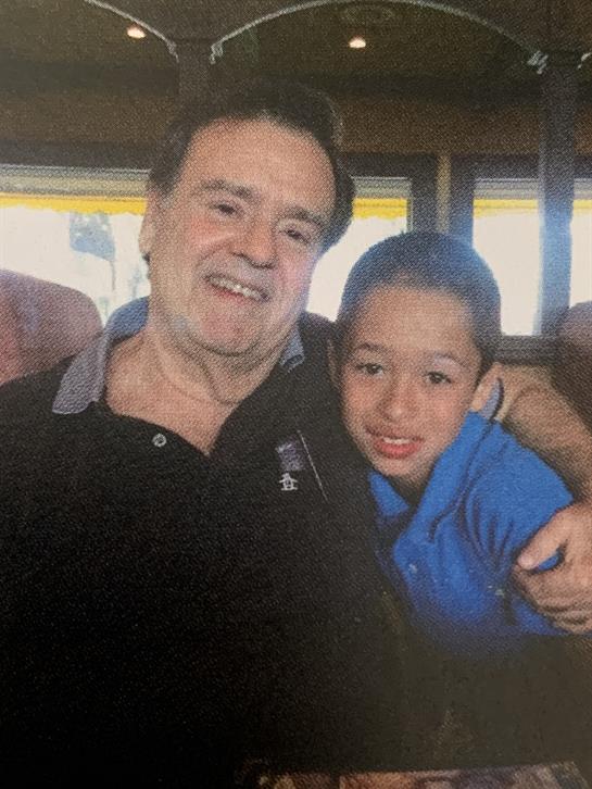 An elderly man embraces his young grandson, both smiling warmly in a relaxed indoor ambiance.