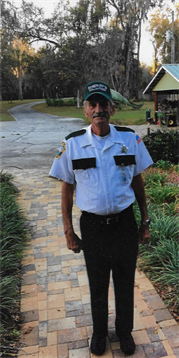 A uniformed security guard stands at the entrance of a suburban neighborhood on a cloudy day.