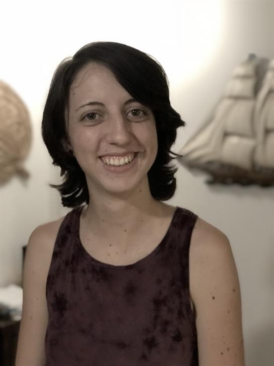 A woman with short dark hair smiles while standing in a warmly decorated room with ship decor.
