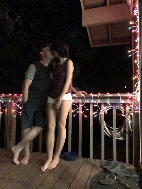Two people embrace and kiss on a wooden deck lit by bright string lights under the night sky.
