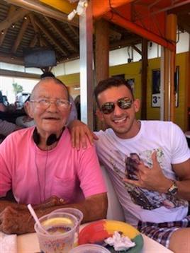 Two men, one elderly and one younger, share laughter and drinks at a vibrant beachside cafe.