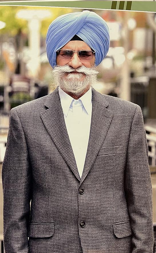 Elderly man in a blue turban and suit walks through a bustling marketplace.