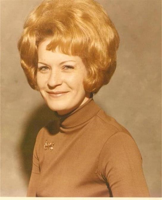 A woman with large curly blonde hair poses confidently in a retro outfit, smiling warmly.