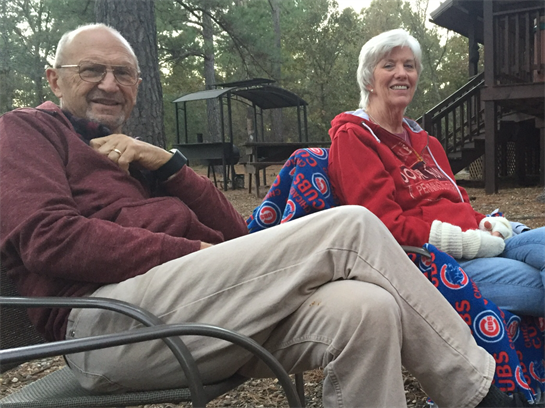 An elderly couple relaxes in chairs, sharing smiles amidst trees in a serene autumn atmosphere.