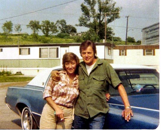 A smiling couple poses together in front of a blue classic car in a trailer park on a sunny day.
