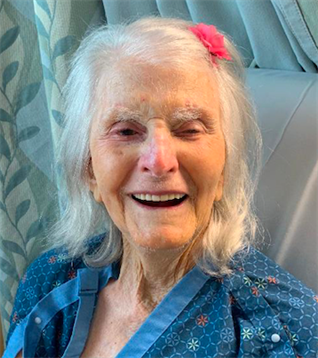 Joyful elderly woman with white hair and a pink flower in her hair smiles in a bright setting.