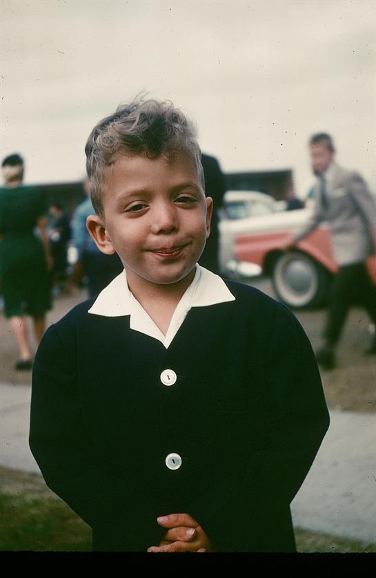 A young boy smiles confidently while surrounded by people and vintage cars at an outdoor gathering.