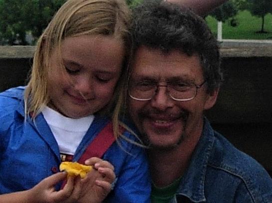 A man and a girl smile together while enjoying a snack in a vibrant park, radiating happiness.