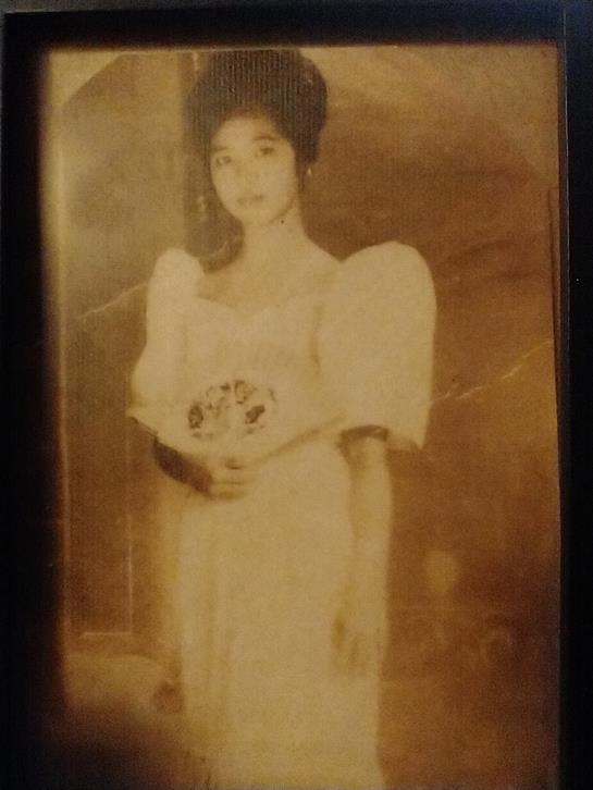 A young woman stands elegantly in a vintage gown, holding flowers amidst a warm background.