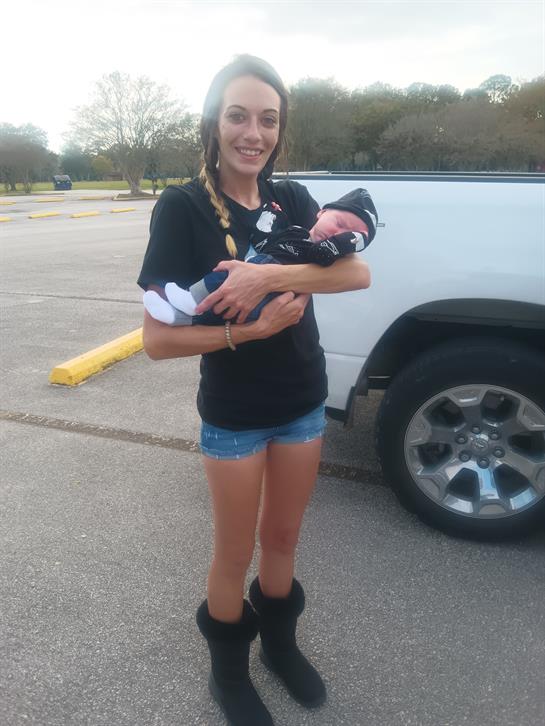 A young woman stands proudly while cradling a newborn baby in her arms outdoors.