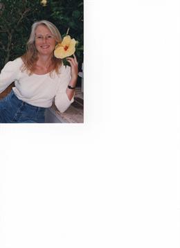 A cheerful woman sits in a garden, showcasing a bright yellow flower while enjoying a sunny day.