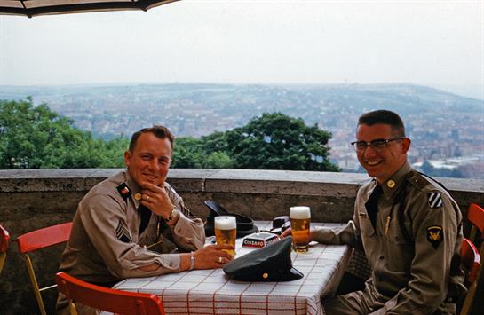 Two smiling men wearing uniforms sit at a table, sharing drinks and enjoying a panoramic view.
