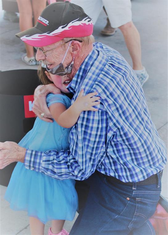 A grandfather in plaid joyfully hugs his granddaughter in a light blue dress.