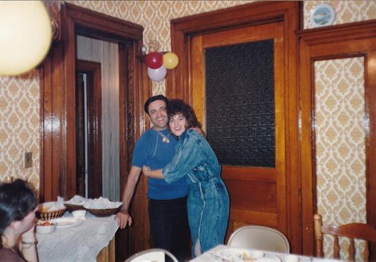 A couple smiles and embraces in a warmly decorated room filled with wooden details.